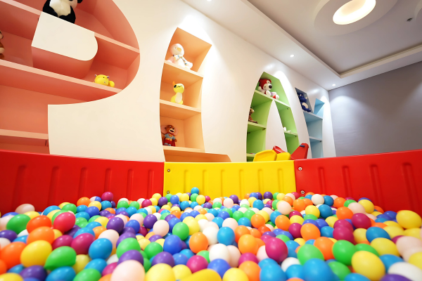 A vibrant and safe children's indoor play area is visible, confirming that these Premium Flats in Guwahati are family-friendly. The room features a large ball pit filled with multi-coloured balls, soft interlocking floor mats, and charming wall-mounted shelves spelling "PLAY" adorned with plush toys. This dedicated, brightly lit space provides a stimulating and secure environment where children can engage in active play, ensuring a comfortable and well-rounded living experience for young families.