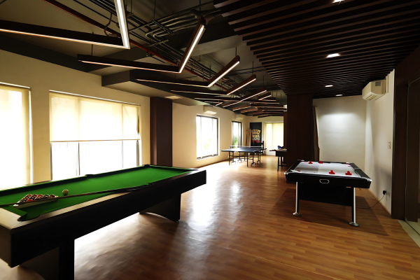 This image showcases a modern, well-lit indoor games room, a fantastic amenity in Premium Flats in Guwahati. The space features a full-sized billiard table, a separate pool table, and a table tennis setup further back. Stylish linear lighting fixtures and a slatted wooden ceiling add to the upscale ambiance. Large windows with blinds ensure ample natural light, creating a perfect retreat for residents to relax and socialize over a friendly game, enhancing a luxury lifestyle.