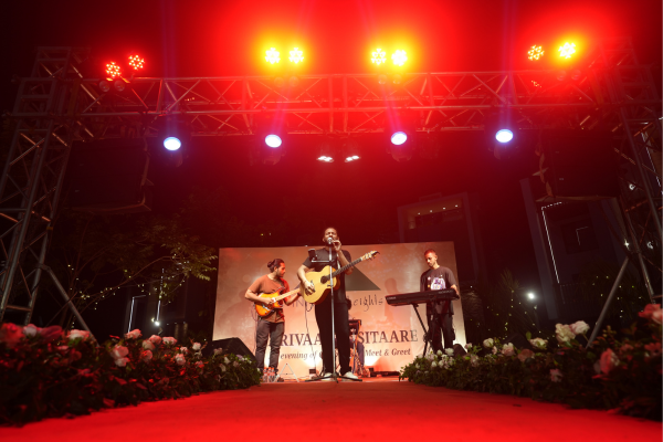 A live band performs on an outdoor stage under bright red and white lights. This musical performance was a major attraction at the Infinity Heights "Parivaar ke Sitaare" evening.