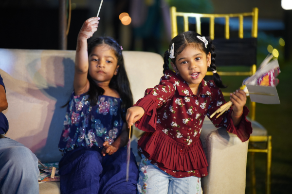 Two young girls sit on a couch, holding up handmade crafts during a festive family event. They are enjoying the celebrations at Infinity Heights.