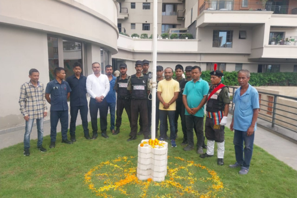 A group of men, including security staff and residents, stand around a decorated flagpole at Infinity Heights, appearing to prepare for or conclude a flag-hoisting ceremony.