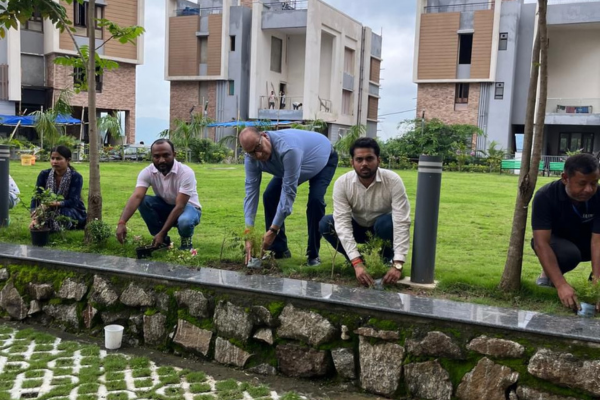 A group of residents and staff at Infinity Heights are gathered on a grassy lawn for a tree-planting activity. One man waters a new sapling with a green watering can, while others look on, participating in the community event.