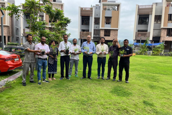 A group of residents and staff at Infinity Heights are gathered on a grassy lawn for a tree-planting activity. One man waters a new sapling with a green watering can, while others look on, participating in the community event.