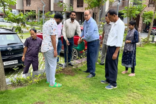 A group of residents and staff at Infinity Heights are gathered on a grassy lawn for a tree-planting activity. One man waters a new sapling with a green watering can, while others look on, participating in the community event.
