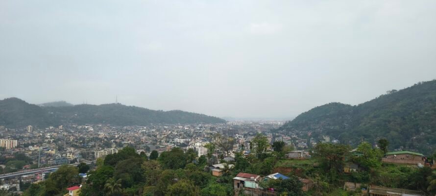 A high-angle panoramic view of Guwahati's urban landscape nestled between lush, green hills under an overcast sky.
