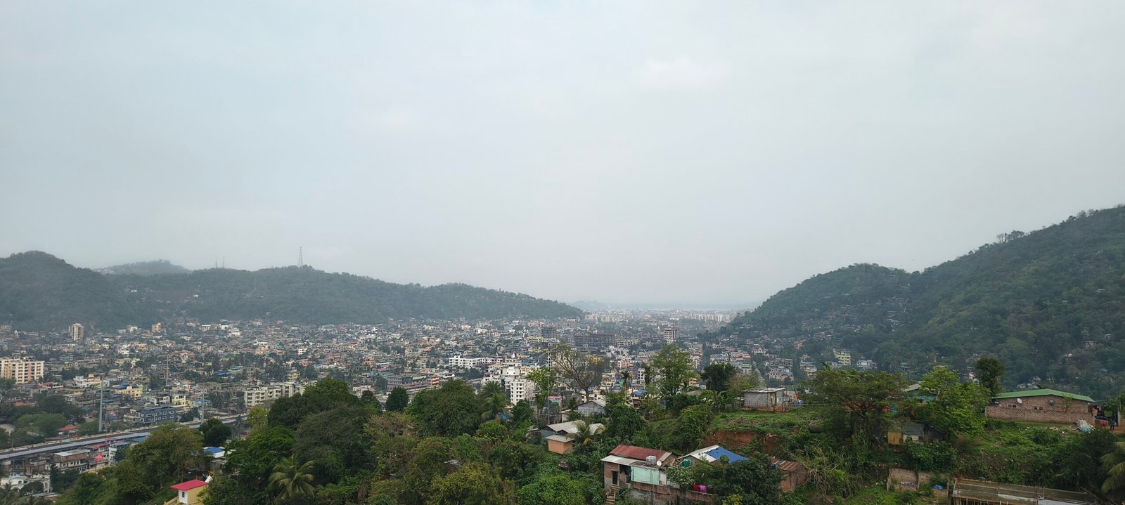 A high-angle panoramic view of Guwahati's urban landscape nestled between lush, green hills under an overcast sky.