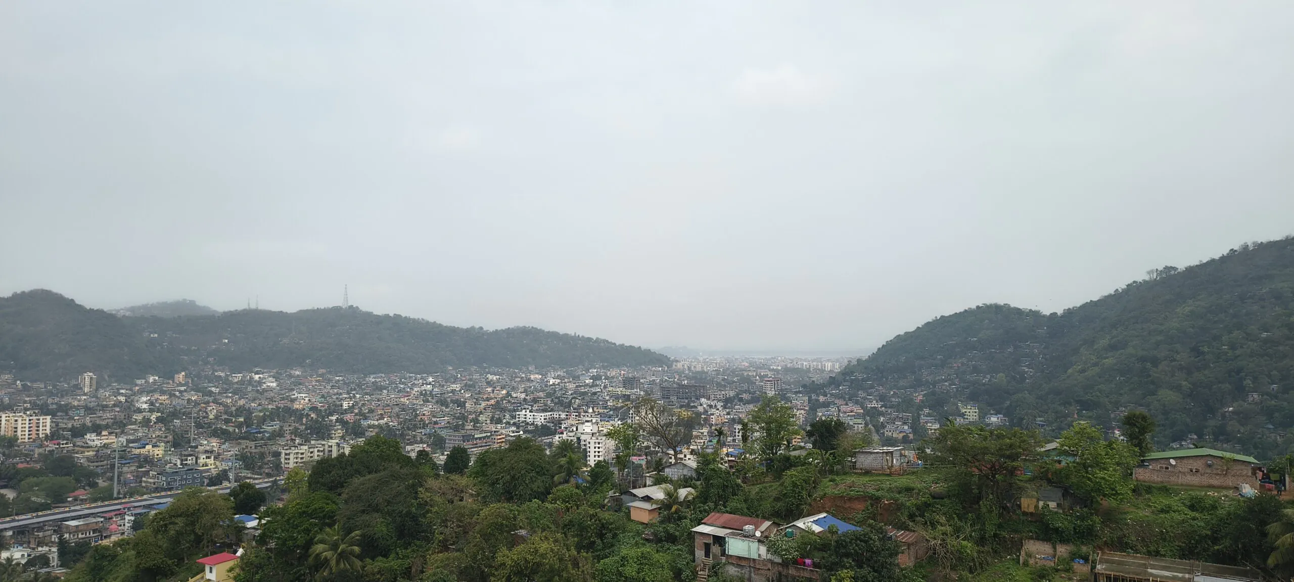 A high-angle panoramic view of Guwahati's urban landscape nestled between lush, green hills under an overcast sky.