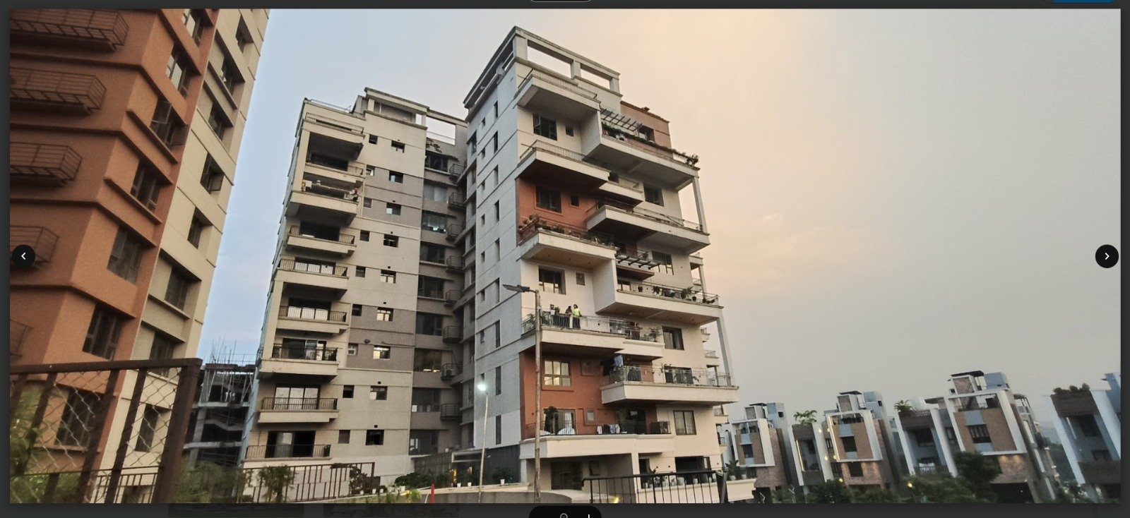A modern, multi-story luxury apartment complex with architectural balconies and brick accents stands under a sunset sky, symbolizing Guwahati's vertical transformation.