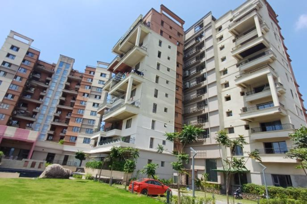A low-angle exterior shot of high-rise luxury apartment towers in Guwahati featuring modern balconies, terra-cotta and off-white facades, surrounded by a manicured green lawn, young trees, and a parked red car under a clear blue sky.