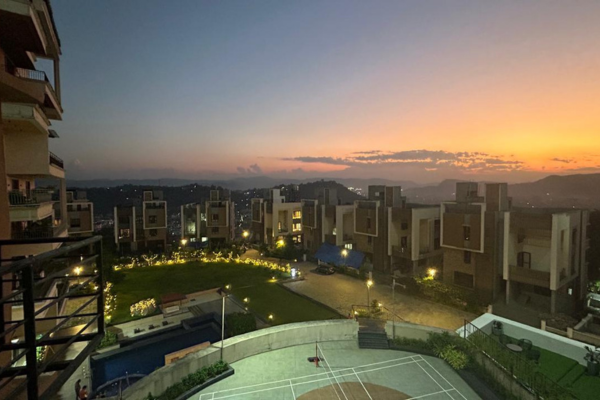 Elevating high-angle dusk view of a residential luxury apartment complex featuring illuminated villas, a badminton court, swimming pool, and central lawn set against a vibrant sunset sky and surrounding hills.