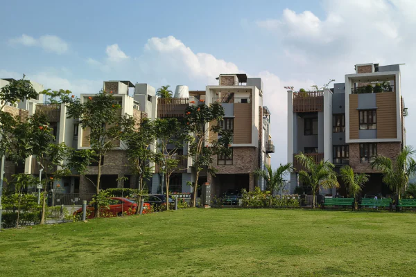 A closer view of modern multi-story townhouses with stone and wood panel exteriors, situated behind a green lawn and young trees under a partly cloudy sky.