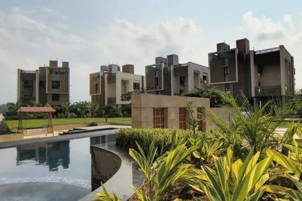 An exterior view of a luxury residential complex featuring modern multistory buildings with wood and white facade accents, a large curved swimming pool, green gardens, and palm trees under a bright sky, showcasing the premium features of upscale apartments in Guwahati.