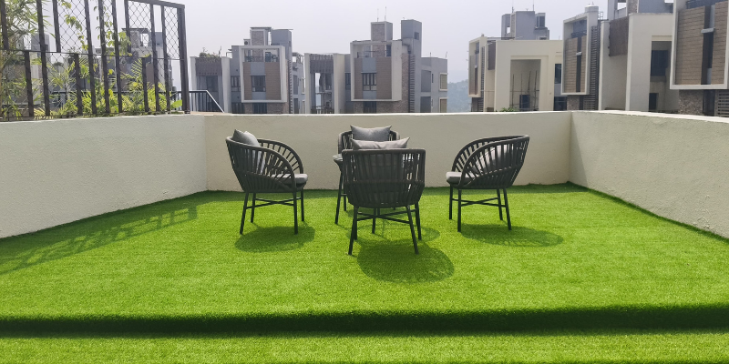 An outdoor rooftop sitting area featuring modern wicker patio chairs arranged on green artificial turf grass, showcasing premium balcony designs ideal for modern apartments in guwahati with residential buildings in the background.