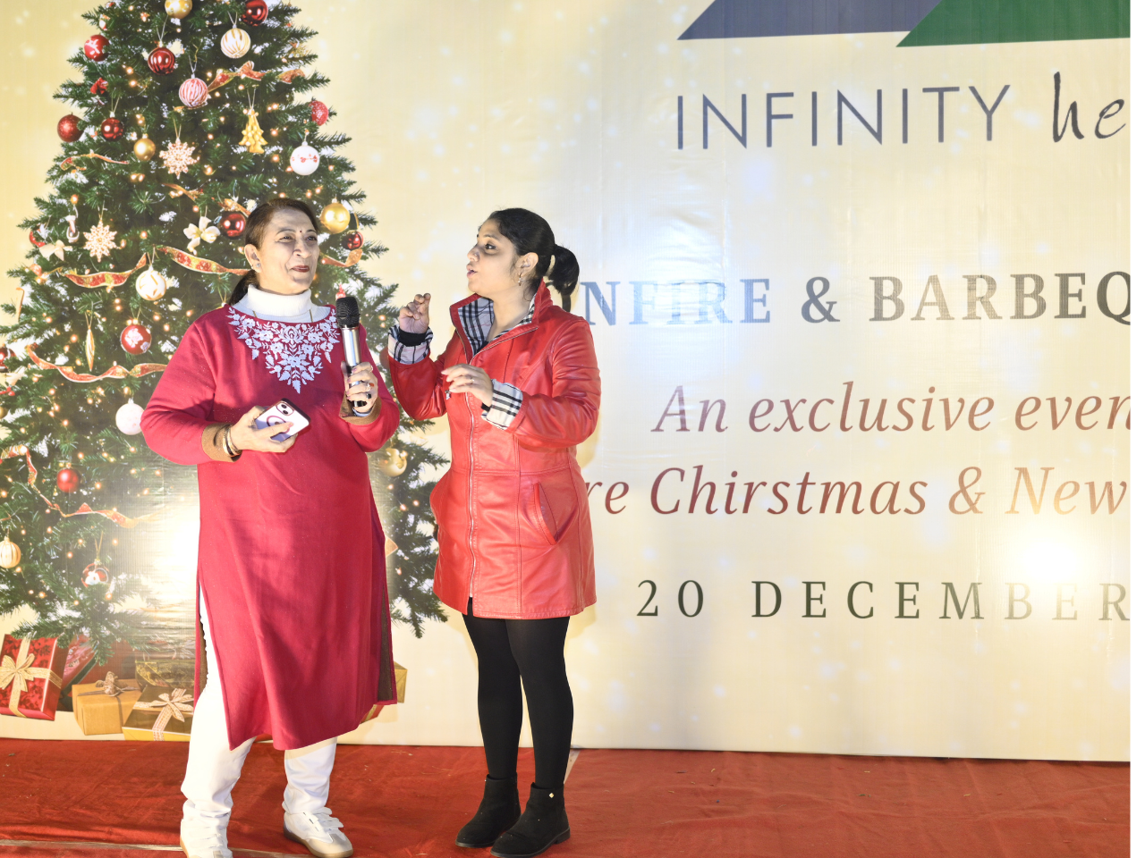 Event hosts speaking in front of the Christmas tree backdrop during the holiday bonfire at an Infinity Heights apartment in Guwahati.