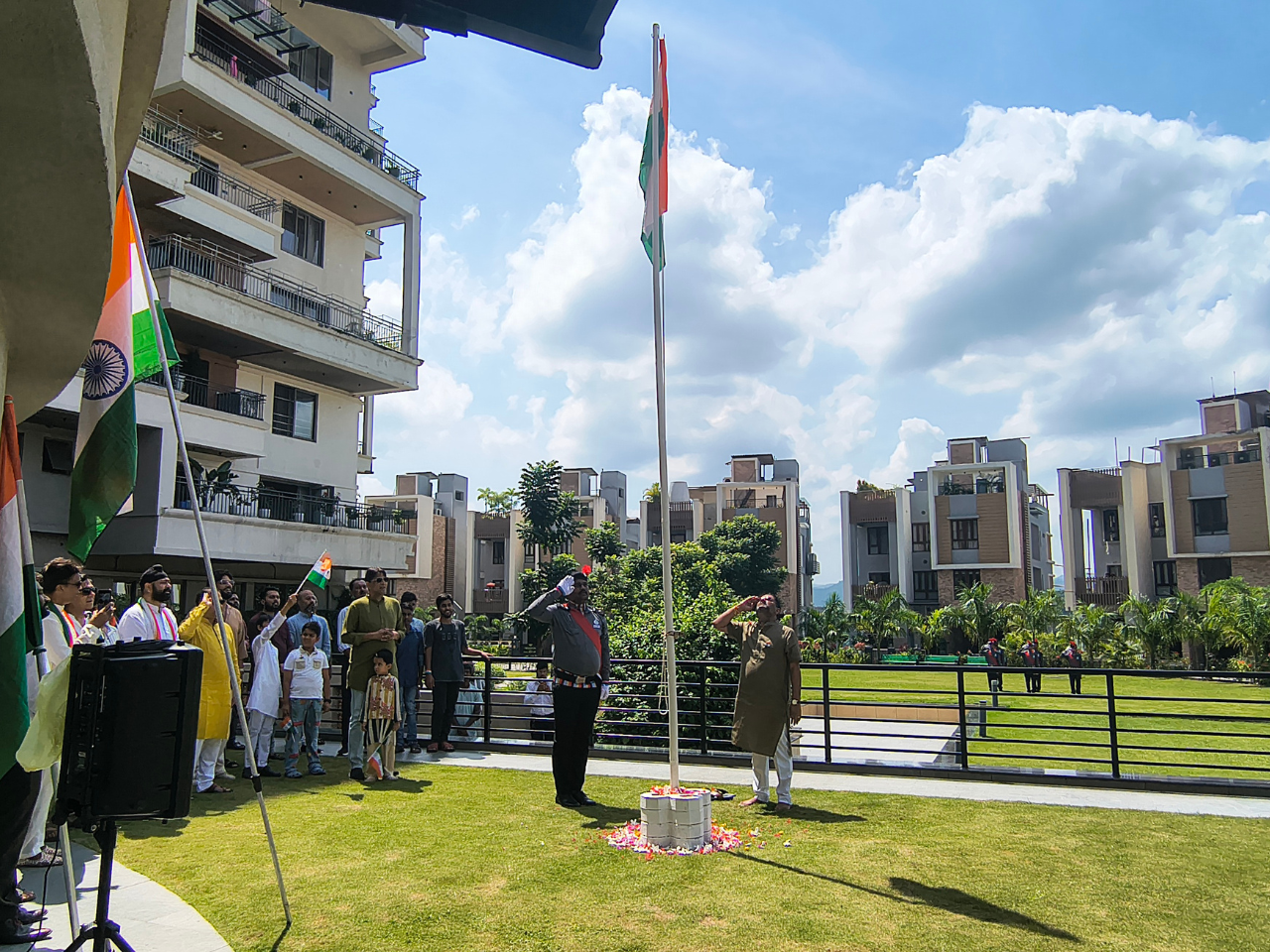 A man and a security guard saluting the hoisted Indian national flag outdoors, surrounded by residents of Infinity Heights.