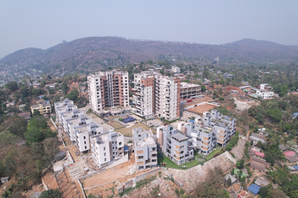 An aerial high-angle view of a modern residential complex featuring high-rise apartment towers and multi-story row houses built into a hillside, surrounded by green hills under a clear sky.