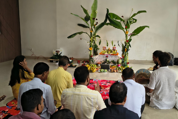 Devotees gathered on floor mats facing a decorated alter framed by banana trees during a traditional puja ritual.