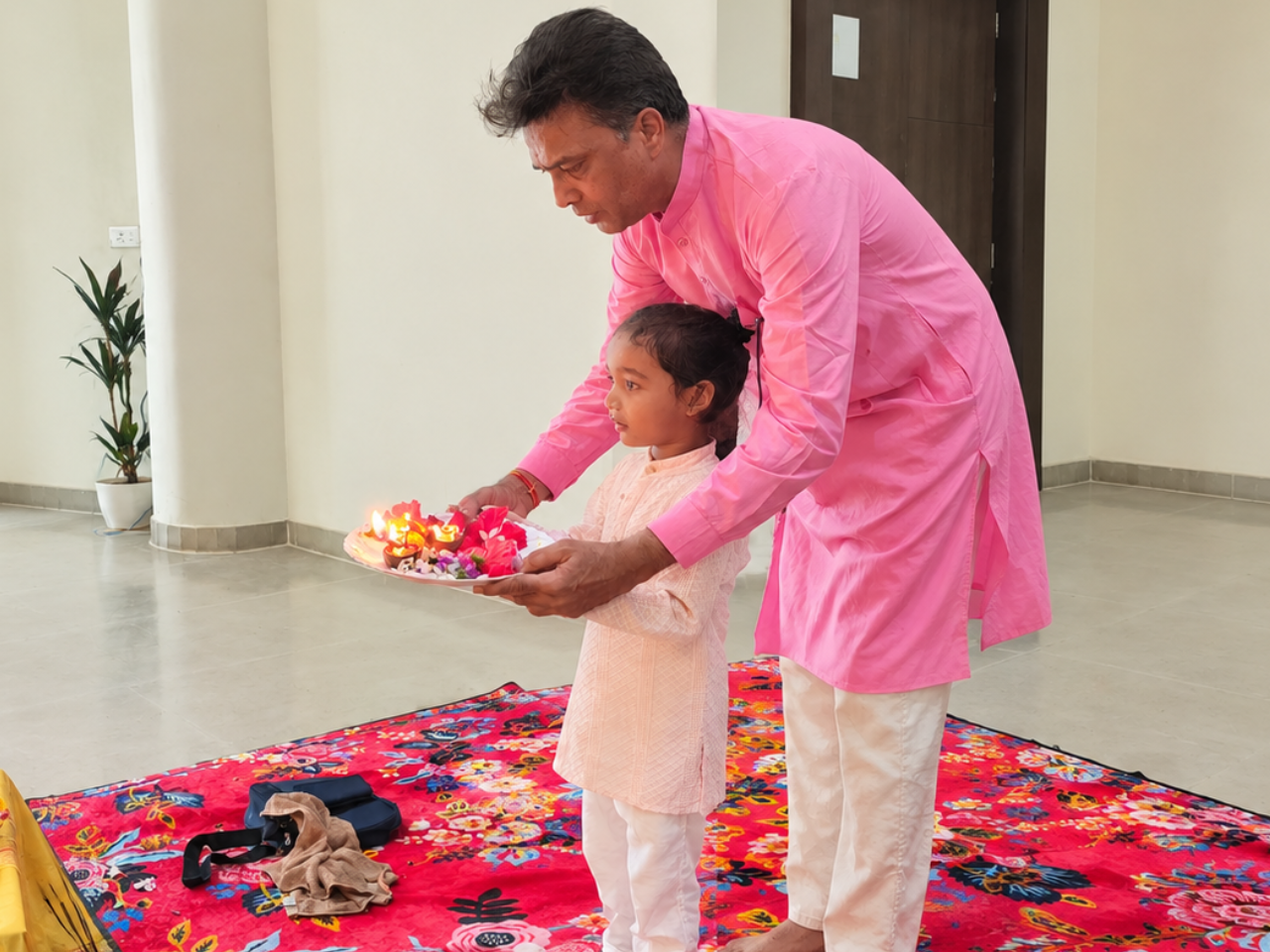 Man in pink kurta guiding a child holding an aarti plate at an altar.